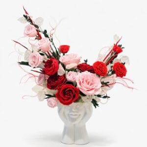 Preserved red and blush pink rose floral arrangement in a white sculptural ceramic face vase on a clean white background