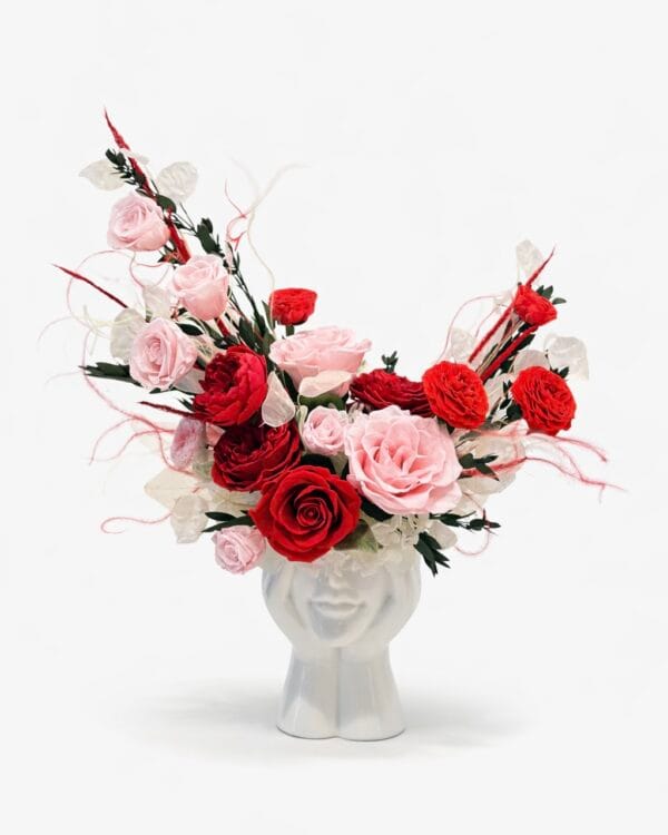 Preserved red and blush pink rose floral arrangement in a white sculptural ceramic face vase on a clean white background
